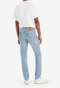 Personne portant un jean bleu clair, un t-shirt blanc et des baskets blanches, vue de dos sur un fond blanc uni.