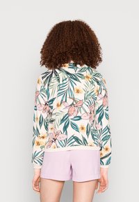 Femme aux cheveux bouclés portant un haut à manches longues à motifs floraux et un short violet clair, debout de dos sur un fond uni.