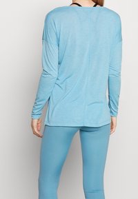 Kvinna som bär en ljusblå långärmad topp och matchande leggings, stående med ryggen mot kameran framför en neutral bakgrund.