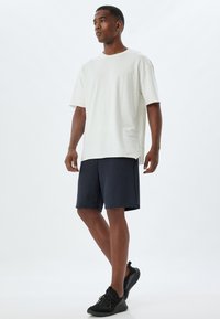Vit bomullströja med kort ärm, parat med mörkblå shorts. Svarta sneakers med strukturerad yta och snören fulländar looken.