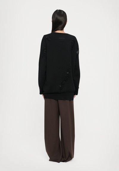 Black oversized sweater with distressed accents, featuring a crew neckline and loose fit. Paired with wide brown trousers in a soft fabric.