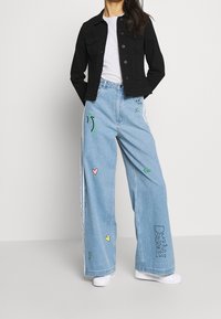 Svart denimjacka, vit t-shirt, vida ljusblå jeans med färgglada broderade mönster, vita sneakers och synliga sidostreifen.