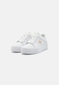 White low-top sneakers with thick soles, perforated detailing, laces, and a gold "CK" logo on the sides and tongues.