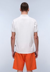Hvid polo t-shirt med korte ærmer; glat tekstur. Sammen med livlige orange shorts. Enkel krave-design; minimal branding detaljer.