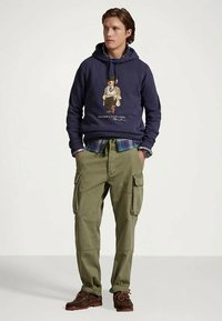 Junger Mann, der einen navy Hoodie mit Bärenmotiv, ein kariertes Hemd, olivgrüne Cargohosen und braune Lederschuhe trägt, steht mit den Händen in den Taschen.