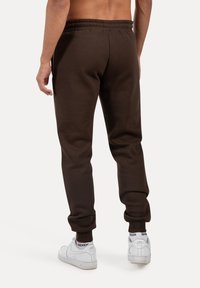 Brune sweatpants med en tapered pasform og elastisk talje, præsenteret sammen med hvide sneakers på en neutral baggrund.