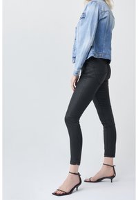 Kvinna iförd en ljusblå denimjacka, svarta skinnyjeans och svarta sandaletter med låg klack, stående i profil mot en enkel bakgrund.