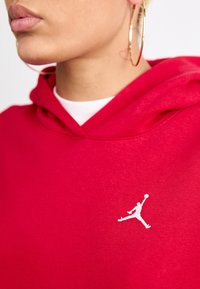 Rød hettegenser med en myk tekstur, med en hvit Jordan-logo på brystet. Hette har et klassisk design med en snorlukking.