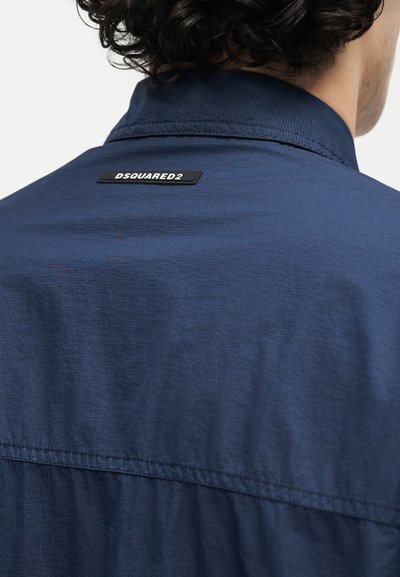 Άνδρας με σκουρόχρωμα σγουρά μαλλιά που φοράει μπλε πουκάμισο ναυτικού χρώματος με μαύρη ορθογώνια ετικέτα DSQUARED2 στο πίσω μέρος του γιακά.