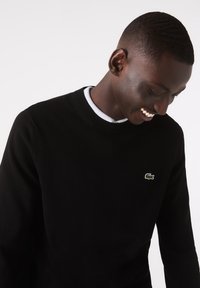 Maglione nero lavorato a maglia con scollo a girocollo e polsini a costine, caratterizzato da un piccolo logo verde Lacoste sul petto sinistro. Texture liscia.
