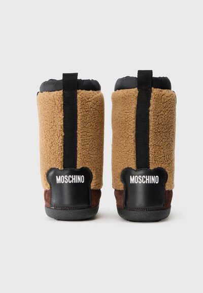 Bottes en shearling marron avec accents noirs, comportant un col rembourré, une languette à l'arrière, et le logo "Moschino" en blanc sur le talon. Semelle texturée.