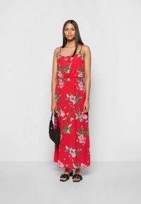 Vestido maxi floral vermelho com alças finas, cintura franzida e folhas verdes. Combinado com uma bolsa tecida preta e sandálias pretas de tiras.