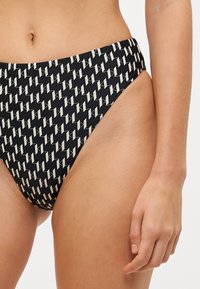 Fekete bikini alsó, magas kivágással, geometrikus fehér mintával. Simító anyagból készült, rugalmas derékkal.