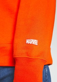 Sudadera naranja con tejido texturizado. Presenta el logo blanco de "MARVEL" en el puño. Costuras visibles y ajuste casual.
