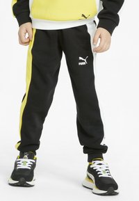 Dziecko noszące czarne, żółte i białe spodnie dresowe Puma oraz dopasowane sneakersy Puma z białymi sznurówkami na białym tle.