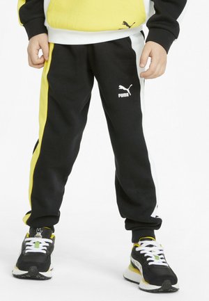 Kind, das schwarze, gelbe und weiße Puma-Sweatpants und passende Puma-Sneaker mit weißen Schnürsenkeln trägt, vor einem weißen Hintergrund.