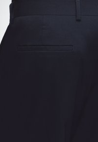 Primo piano della tasca posteriore di pantaloni formali blu scuro con passante per cintura visibile in vita.