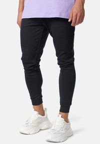 Parte inferior de una persona que lleva pantalones jogger negros, zapatillas deportivas blancas y una camiseta de color púrpura claro, todo contra un fondo blanco.