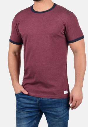 Man wearing a maroon short-sleeve t-shirt with black trim on collar and sleeves, paired with blue jeans, standing with one hand in pocket.