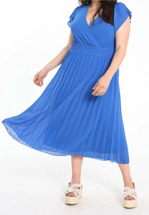Mujer con vestido midi plisado azul con escote en V y mangas casquillo, combinado con sandalias de plataforma beige, de pie sobre fondo blanco.