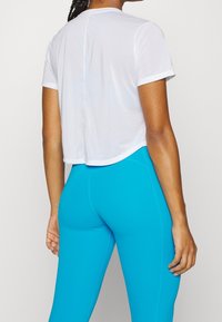 Camiseta blanca corta con mangas cortas y dobladillo redondeado, combinada con leggings azul brillante. Textura suave y diseño atlético.