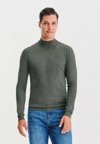 Ciemnozielony sweter z golfem, wykonany z gładkiego materiału, z długimi rękawami i dopasowanym krojem, w połączeniu z niebieskimi dżinsami.