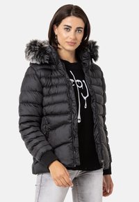 Schwarze Pufferjacke mit Kapuze aus Kunstfell, gestepptem Muster, Reißverschlusstaschen und Rippbündchen, getragen über einem schwarzen Pullover mit Grafikdesign.