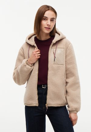 Kvinde iført beige fleecejakke med hætte og bordeaux skjorte, stående foran en ensfarvet baggrund.