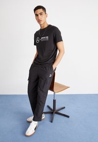 Zwarte T-shirt met wit logo, gecombineerd met zwarte cargobroek met zakken. Model draagt witte sneakers met beige accenten, tegen een effen achtergrond.