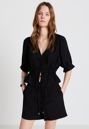 Frau mit welligem hellbraunem Haar, die ein schwarzes V-Ausschnitt-Oberteil mit taillierter Passform und passende schwarze Shorts trägt, silbernes Manschettenarmband am linken Handgelenk.