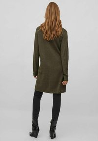 Abito maglione verde oliva con maniche lunghe, polsini e orlo a costine. Indossato sopra jeans skinny neri e stivaletti con tacco. Texture liscia.