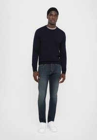 Maglione lavorato a maglia di colore blu navy con scollo rotondo, abbinato a jeans slim-fit blu scuro e sneaker bianche, su uno sfondo bianco semplice.