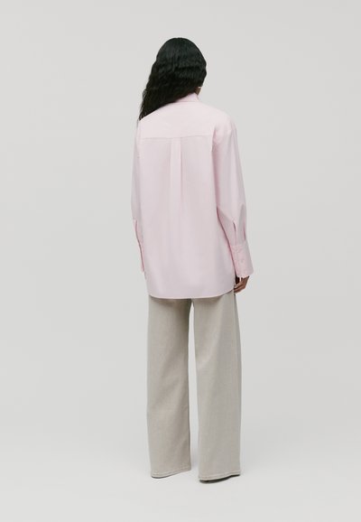 Camicetta rosa chiaro a maniche lunghe con dettaglio a yoke sul retro, vestibilità comoda e polsini a bottone, abbinata a pantaloni grigi chiari a gamba larga.