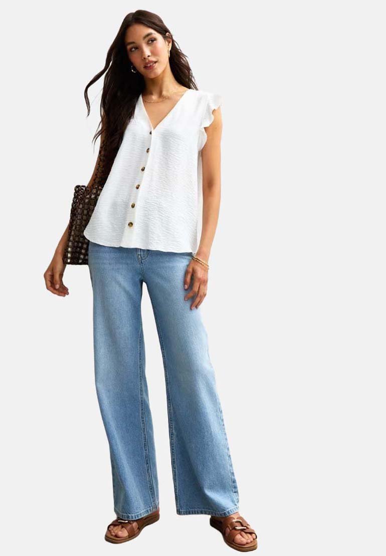 Vit, texturerad blus med V-ringning och volangärmar samt guldfärgade knappar, kombinerad med ljusblå vida jeans och bruna sandaler.