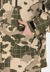 Camouflagejakke i beige og grøn, med et rutemønster, en sidelomme og en struktureret overflade med velcro-lukning ved manchetten.