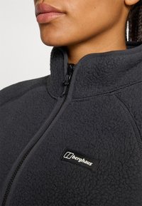 Zbliżenie na szyję i szczękę osoby w ciemnej polarowej kurtce z zamkiem, z czarnym logo "Berghaus" na piersi.