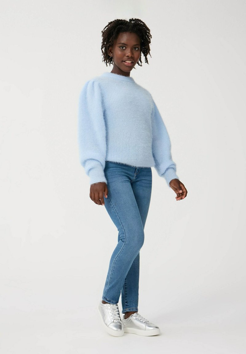 Jasnoniebieski puszysty sweter z bufiastymi rękawami, w zestawieniu z dopasowanymi ciemnoniebieskimi dżinsami i srebrnymi trampkami. Gładkie tło.