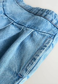 Shorts en denim bleu clair avec un détail plissé, une taille élastique et des surpiqûres contrastantes le long des coutures. Comprend une poche latérale.