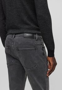 Jeans de denim gris oscuro con una textura suave, presentan un parche de cuero negro con marca en la cinturilla y dos bolsillos traseros.