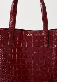 Sac fourre-tout bordeaux avec une texture brillante en relief crocodile. Il dispose de deux poignées solides et d'un logo doré. Fini lisse et brillant.