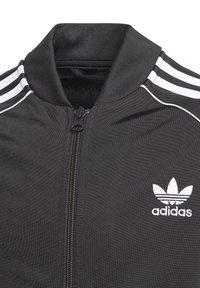 Musta track jacket, jossa on vetoketjullinen etuosa, ribbauskaulus ja kolme valkoista raitaa hartioilla. Adidas-logo valkoisena vasemmalla puolella.