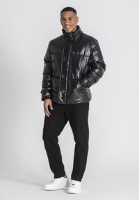 Giacca nera imbottita con colletto alto, texture lucida e cuciture a vista. Indossata sopra pantaloni neri e sneakers bianche, in uno stile casual.
