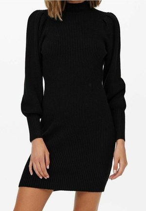 Femme portant une mini robe en maille côtelée noire avec des manches longues bouffantes et un col montant, vue des épaules jusqu'au milieu des cuisses.