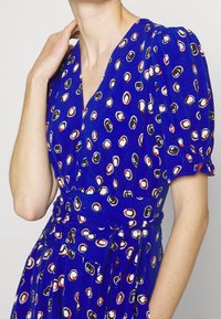 Blue dress with a v-neckline, short puff sleeves, and a fitted waist. Features a pattern of small white, black, and red circles on the fabric.