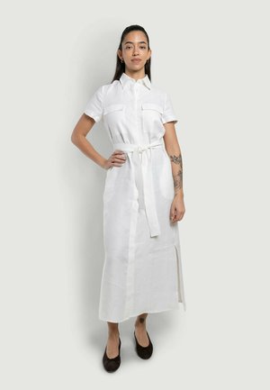 Donna con lunghi capelli scuri, indossa un abito camicia bianco con cintura, maniche corte e ballerine nere, in piedi con una mano sui fianchi davanti a uno sfondo semplice.