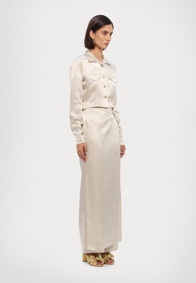 Cream-colored, silky two-piece outfit features a cropped button-up jacket with pockets and a long, draped skirt with a smooth texture.
