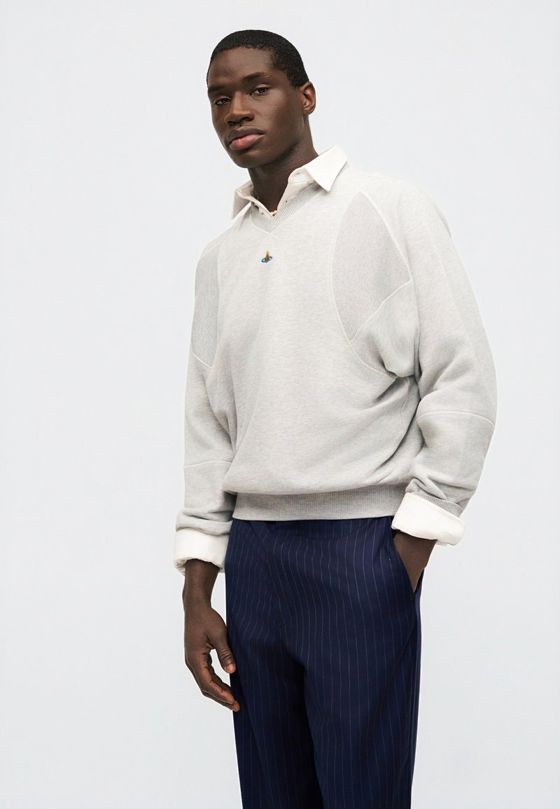 Giovane uomo che indossa un maglione grigio chiaro sopra una camicia bianca con colletto e pantaloni blu navy a righe sottili, in piedi con una mano in tasca.