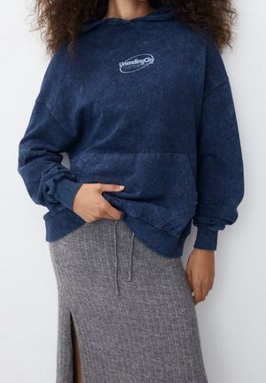 Woman wearing a navy blue hoodie with "Unending City" text and a gray ribbed knit skirt with a front slit.