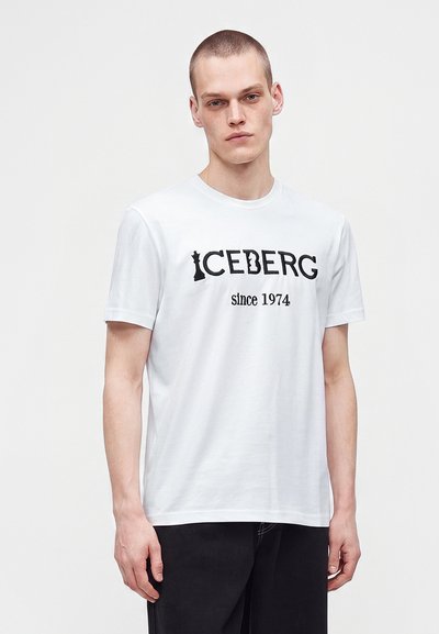 Млад мъж с къса коса, носещ бяла тениска с отпечатан в черно надпис "ICEBERG since 1974", съчетана с черен панталон на едноцветен фон.