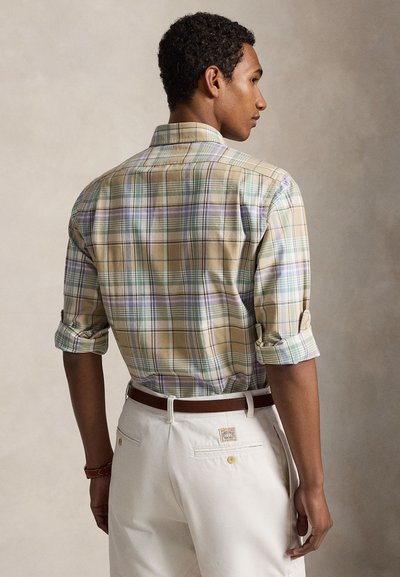 Homme portant une chemise à carreaux verte, beige et violette avec manches retroussées, pantalon blanc et ceinture marron, debout devant un fond uni.